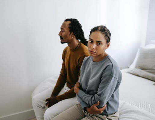 Man and woman sitting on edge of made bed looking unhappy.