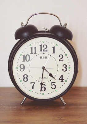 An analog clock sitting on a desk. Has two bells on top that act as an alarm. The image represents TimeTemplates.com.